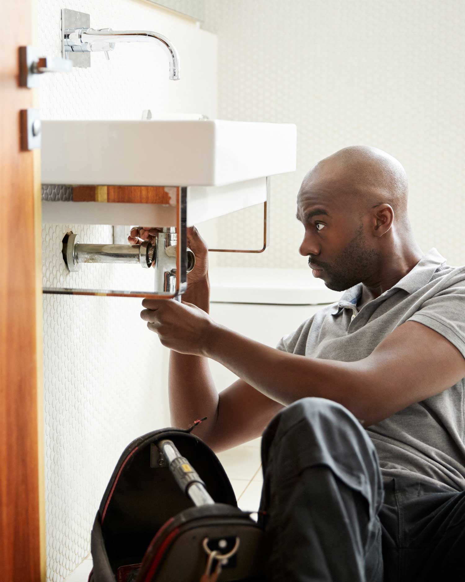 Young black male plumber sitting on the floor fixing a bathroom sink