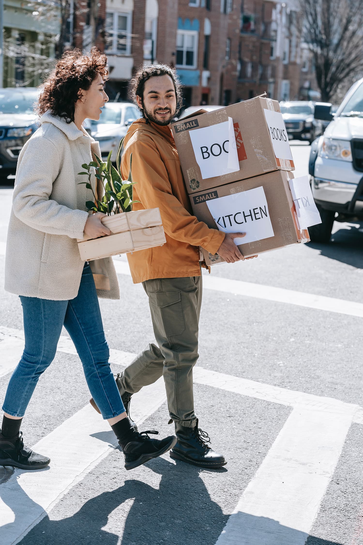 Couple Walking Along The Street Carrying Boxes And Plants